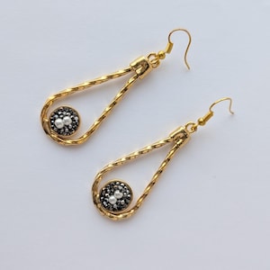 May include: A pair of gold-toned drop earrings featuring a twisted teardrop-shaped frame. Each earring contains a circular charm at the base, encrusted with small dark gray crystals and two small white spherical beads in the center, with hook wires.