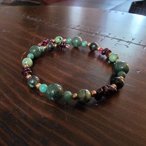 May include: A wrap bracelet featuring a mix of round beads in shades of forest green, turquoise, and deep burgundy. The beads are interspersed with small metallic gold-toned spacers, creating a circular design with varying bead sizes and textures.
