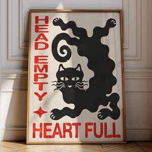May include: A rectangular framed wall art print featuring a stylised black cat with a wavy, elongated body on a cream background. The text "HEAD EMPTY" is printed in red vertically on the left, and "HEART FULL" is printed in red at the bottom.