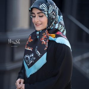 May include: A black headscarf with a butterfly pattern in shades of blue, white, and orange. The scarf has a light blue border and features the text "Mesh" and "Maya Shams". The scarf is worn with a black dress.