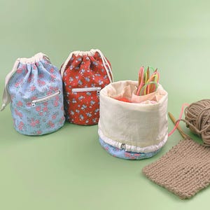 Peut inclure: Deux sacs cylindriques en tissu à cordon avec des motifs floraux bleu clair et rouge, chacun doté d'une poche zippée frontale. Un troisième organisateur en tissu ouvert contient des aiguilles à tricoter, une pelote de laine brune et un échantillon.