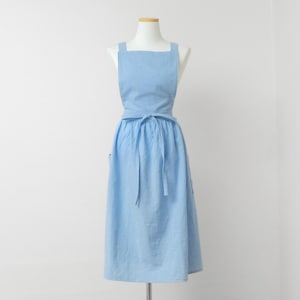 May include: A light blue apron featuring a square bib, wide shoulder straps, and a gathered skirt that falls to the mid-calf. The apron includes a matching fabric tie at the waist, which is knotted in the center, and side pockets on the skirt.