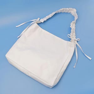 May include: A solid white hobo-style shoulder bag featuring a gathered, scrunchie-like strap design. The bag includes thin fabric ties at both ends of the strap where it attaches to the main body, providing a simple and clean aesthetic.