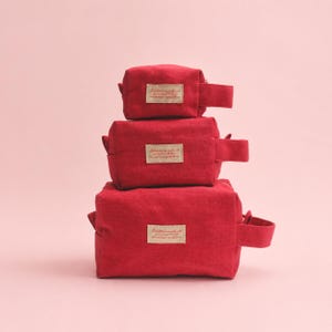 Può includere: Tre astucci rettangolari rossi con cerniera impilati verticalmente, ognuno dotato di una maniglia laterale e di una piccola etichetta rettangolare beige sul davanti con il testo stampato: Each little one made with you in mind. To show how much you enjoy to me.