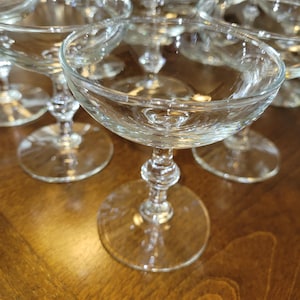 May include: A collection of clear glass coupe glasses, each featuring a wide, shallow bowl, a slender stem with a decorative central knob, and a circular base. The glassware is transparent and designed for serving beverages in a classic style.