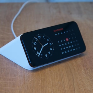 Può includere: Un moderno orologio digitale con display nero che mostra l'ora e un calendario di novembre. L'orologio ha una base bianca angolata e un cavo bianco. Il quadrante mostra l'ora come le 10:07.