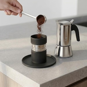 Könnte beinhalten: Ein Mokkakocher aus Edelstahl mit schwarzem Griff steht neben einem schwarzen zylindrischen Kaffeefilterkorb auf einem passenden schwarzen runden Sockel. Ein silberner Löffel schüttet dunkelbraunes Kaffeepulver in den Filterkorb.