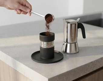 Peut inclure: Une cafetière italienne en acier inoxydable avec une poignée noire est placée à côté d'un panier filtre à café cylindrique noir sur une base circulaire noire assortie. Une cuillère argentée verse du café moulu brun foncé dans le filtre.