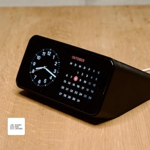 Può includere: Un orologio digitale nero con un display del calendario che mostra il mese di ottobre. Il quadrante mostra l'ora con lancette e numeri bianchi. L'orologio è su una superficie di legno.