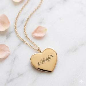 May include: A gold-colored heart-shaped pendant necklace with a delicate chain. The pendant is engraved with the word "Abuela" in cursive script, accented with small floral designs. The necklace rests on a marble surface with scattered pink petals.