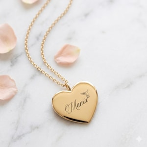 May include: A gold-colored heart-shaped pendant necklace. The pendant has the word "Mama" engraved in cursive, along with a hummingbird illustration. The necklace chain is also gold-colored, and the background features scattered pink flower petals.