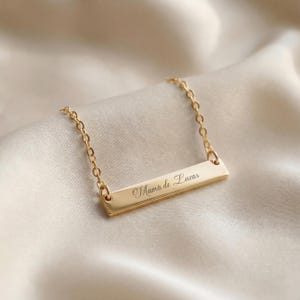 May include: A gold-colored bar necklace with a delicate chain. The bar is engraved with the words "Mamá de Lucas" in cursive script. The necklace is displayed on a cream-colored, satin-like fabric.