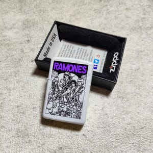 Pode incluir: Um isqueiro Zippo branco com um gr&aacute;fico a preto e branco da banda Ramones, com o nome da banda em roxo. O isqueiro est&aacute; numa caixa preta com o log&oacute;tipo Zippo e "Made in USA" impresso.