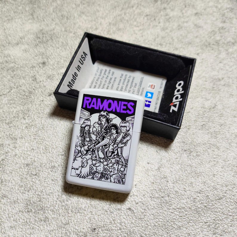 Pode incluir: Um isqueiro Zippo branco com um gr&aacute;fico a preto e branco da banda Ramones, com o nome da banda em roxo. O isqueiro est&aacute; numa caixa preta com o log&oacute;tipo Zippo e "Made in USA" impresso.