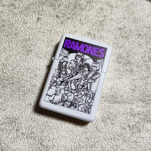 Pode incluir: Isqueiro branco com uma ilustra&ccedil;&atilde;o a preto e branco de uma banda. A palavra "RAMONES" est&aacute; impressa em roxo na parte superior do isqueiro.