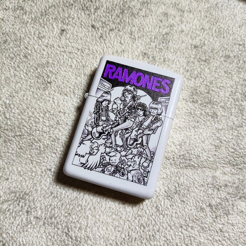 Pode incluir: Isqueiro branco com uma ilustra&ccedil;&atilde;o a preto e branco de uma banda. A palavra "RAMONES" est&aacute; impressa em roxo na parte superior do isqueiro.