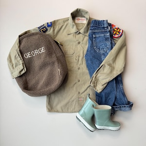 May include: A tan button-up shirt, blue jeans, and a brown sherpa backpack with the name "GEORGE" embroidered in white. The outfit includes light green rain boots with white soles. Patches are sewn on the shirt.