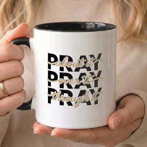 Peut inclure: Mug en céramique blanche avec intérieur et anse noirs. Le mug présente les mots "PRAY about it, PRAY over it, PRAY through it" dans un design empilé. Le texte est en noir et or.