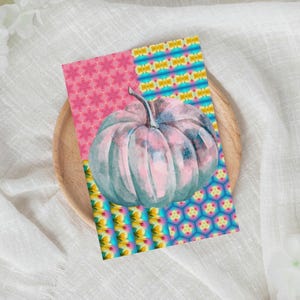 以下が含まれることがあります： 水彩画風のグリーティングカード。水色とピンクのかぼちゃが描かれています。カードの左側にはピンクの花柄、右側にはカラフルな幾何学模様があります。カードは木製のプレートの上に置かれています。