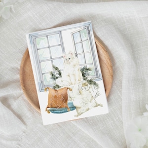 May include: A watercolour illustration of a fluffy white cat in front of a window. The cat sits on a stack of pillows, with a brown and blue cushion in the foreground. The window is open, and there are green sprigs of pine.