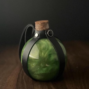 May include: A round glass bottle filled with a shimmering green liquid, secured with a black leather strap and a cork stopper. The bottle has a handle and is set against a dark background.