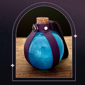 May include: A round, blue glass bottle with a cork stopper and a purple leather strap. The bottle is filled with a shimmering, light blue liquid. The leather strap is secured with silver-colored rivets and a white button.