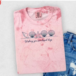 May include: A pink tie-dye t-shirt featuring a dark blue graphic of four seashells and a starfish arranged in a row above the text "Wishing you sun-kissed days." The shirt is paired with blue denim jeans that have distressed, ripped details.