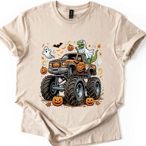 May include: A beige short-sleeve t-shirt featuring a graphic of an orange monster truck with large tires. The truck is decorated with jack-o'-lanterns and carries a green dinosaur wearing a white ghost sheet, surrounded by bats and falling leaves.