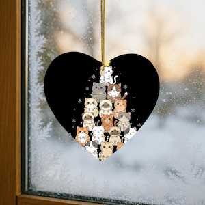 May include: Heart-shaped Christmas ornament featuring a black background with a tree made of cartoon cats. The cats are various colors and patterns, with some wearing string lights. A gold string is attached for hanging.