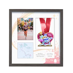 May include: Framed RunDisney Half Marathon memorabilia. Includes a photo of a runner, a race map, and a medal with a pink ribbon. The medal features cartoon characters and the text "RunDisney Marathon Weekend". The frame is dark gray.