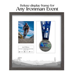 Op de afbeelding: Een grijze display frame met een witte passe-partout, met een Ironman-evenement. De lijst bevat een foto van een fietser, een medaille en racedetails. Tekst luidt "Deluxe display frame for Any Ironman Event."