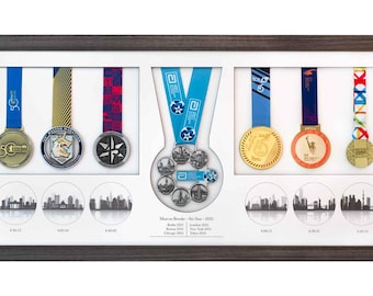 Puede incluir: Una exhibición enmarcada de medallas de maratón con cintas azules, doradas y rojas. Las medallas están en un marco gris oscuro con un fondo blanco. Debajo de las medallas hay siluetas de horizontes urbanos.
