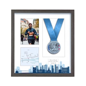 Op de afbeelding: Een ingelijste display met een hardloper, een marathonmedaille en een routekaart. De medaille heeft een blauw lint en de tekst "Detroit Free Press Marathon". De lijst bevat de tekst "Marcus Brooke, October 19th 2025, 5:22:14".