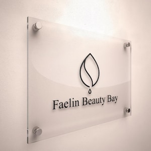 以下が含まれることがあります： 黒い文字で「Faelin Beauty Bay」と書かれた透明なアクリルサイン。サインには、水滴が付いた黒い葉のロゴが付いています。サインは、銀色の金具で白い壁に取り付けられています。