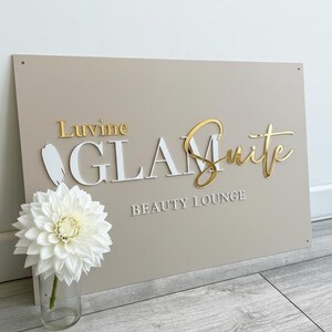 May include: A beige sign with gold and white lettering that reads "Luvine GLAM Suite BEAUTY LOUNGE." A white flower in a glass vase is in the foreground. The sign has a modern, elegant design.