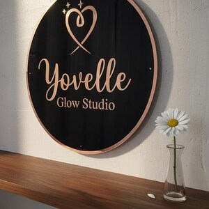 May include: A round black sign with a rose gold border and lettering that reads "Yovelle Glow Studio." The sign features a heart and star design. A small glass vase with a daisy sits on a wooden shelf below the sign.