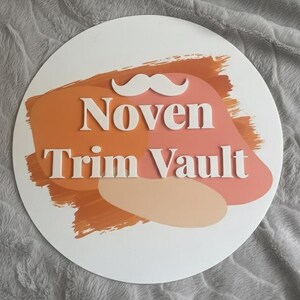 May include: A white, circular sign with the text "Noven Trim Vault" in white, bold letters. The sign features a white mustache graphic above the text. The background has orange, peach, and pink brushstroke designs.
