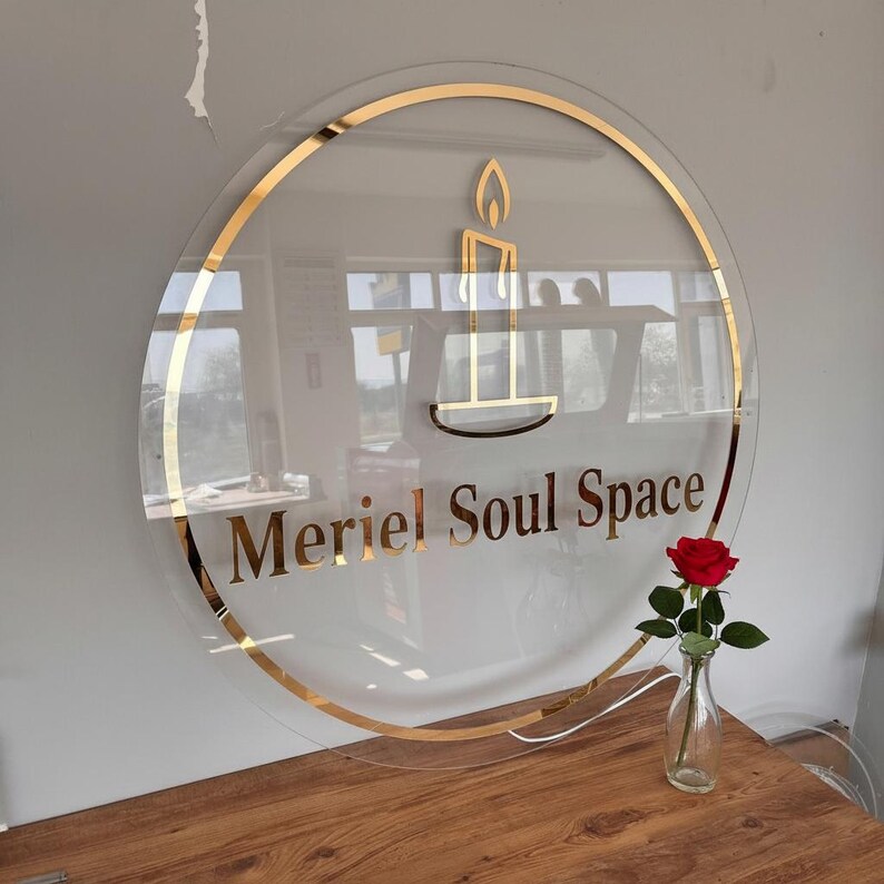 May include: A clear, circular sign with gold trim and lettering that reads "Meriel Soul Space." The sign features a gold candle graphic. A red rose in a small glass vase sits on a wooden surface below the sign.