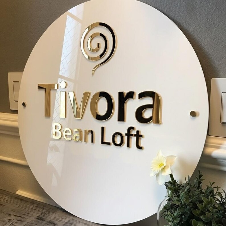 May include: A white, round sign with the words "Tivora Bean Loft" in gold and black lettering. A gold swirl design is at the top. The sign is mounted on a wall with a small flower and greenery in the foreground.