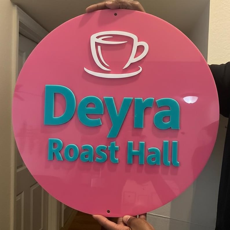 May include: A round, pink sign with the words "Deyra Roast Hall" in teal lettering. A white coffee cup logo is at the top. The sign is held up by a person.