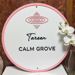 May include: A round white sign with a pink border and a pink embossed design at the top. The sign reads "Tareen CALM GROVE" in black lettering. A vase of white orchids is to the right of the sign.