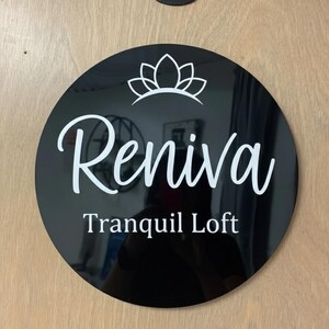 May include: A round, black sign with the name "Reniva" in white script, topped with a white floral design. Below the name, the text "Tranquil Loft" is displayed in a clean, sans-serif font. The sign is mounted on a light-colored wooden surface.