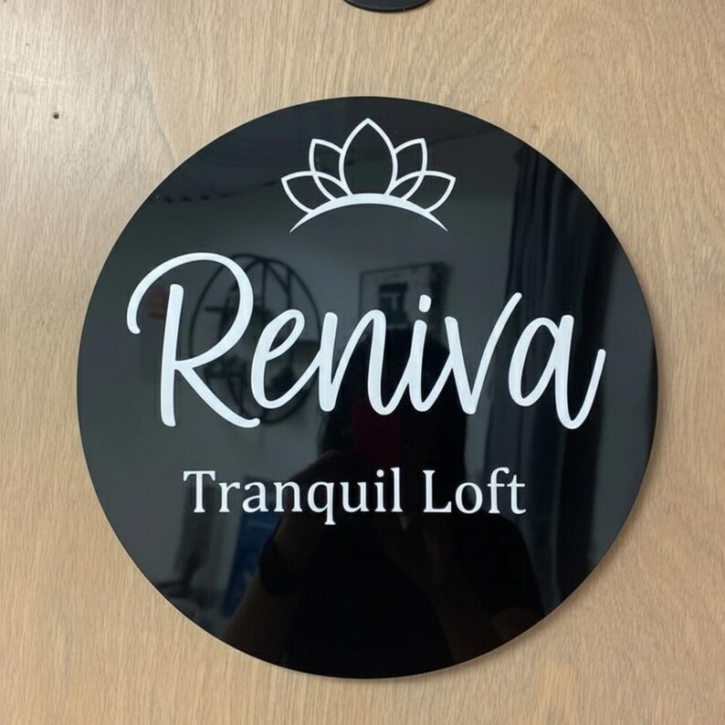 May include: A round, black sign with the name "Reniva" in white script, topped with a white floral design. Below the name, the text "Tranquil Loft" is displayed in a clean, sans-serif font. The sign is mounted on a light-colored wooden surface.
