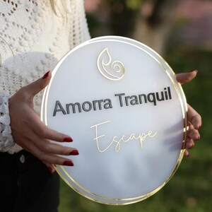 May include: A round, white sign with gold trim and the words "Amorra Tranquil Escape" in black and gold lettering. A gold leaf design is at the top. The sign is held by a person.