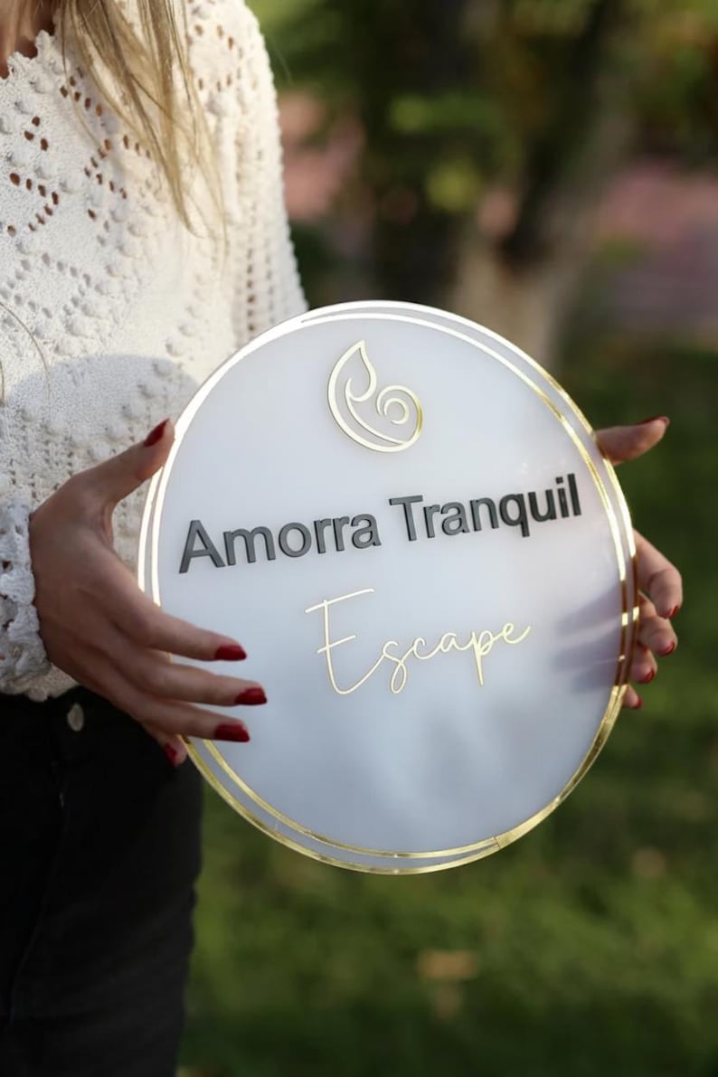 May include: A round, white sign with gold trim and the words "Amorra Tranquil Escape" in black and gold lettering. A gold leaf design is at the top. The sign is held by a person.