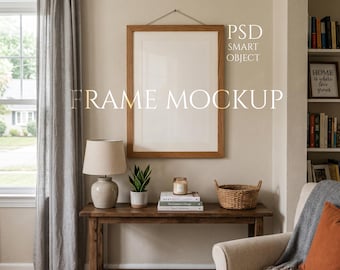 Könnte beinhalten: Ein Holzrahmen-Mockup mit dem Text "FRAME MOCKUP" und "PSD SMART OBJECT". Der Rahmen hängt über einem Holztisch mit Lampe, Pflanze, Büchern, Kerze und Korb. Eine gemütliche Wohnzimmereinrichtung.