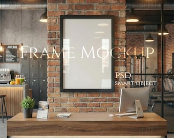 Könnte beinhalten: Ein schwarzer gerahmter Poster-Mockup mit dem Text "FRAME MOCKUP" an einer Backsteinmauer. Ein Holztisch mit Computer, Pflanze und Broschüren befindet sich vor der Wand. Die Szene spielt in einem Fitnessstudio.