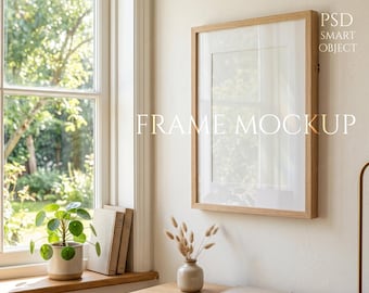 Könnte beinhalten: Ein Holzrahmen-Mockup mit weißem Passepartout, das an einer beigefarbenen Wand hängt. Der Text "FRAME MOCKUP" ist auf dem Rahmen zu sehen. Ein Fenster mit Blick auf einen grünen Garten ist sichtbar.