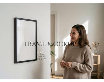 Könnte beinhalten: Ein schwarzer rechteckiger Rahmen-Mockup hängt an einer weißen Wand. Der Rahmen ist leer, mit dem Text "FRAME MOCKUP" und "PSD SMART OBJECT" im Bild. Eine Person in einem beigen Pullover ist im Hintergrund.