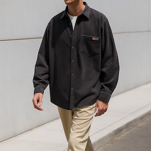 Puede incluir: Una camisa de pana gris oscuro con botones, bolsillo en el pecho y una pequeña etiqueta marrón. La camisa tiene mangas largas y un corte holgado. El modelo lleva pantalones color caqui.